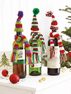 3 wine bottles sitting on drawers. Each bottle has a different coloured scarf with matching bobble hat. Green, purple and red stripes; red, white and a sparkle mix; red, green and white. The scarf has tassels and the hat is topped with a mini pom pom