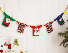 Load image into Gallery viewer, A Christmas garland - yellow rope with mini Christmas jumpers spaced along it. You can see a green sweater with a Santa motif, a red one with a snowman and teal with elf. The other two are knitted in green, white and red self-striping glitz yarn