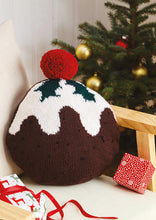 Load image into Gallery viewer, Fun Christmas pudding cushion shown on a grey armchair. The main section is circular and knitted in dark brown DK yarn. The white top is knitted to look like the topping is running down the pudding. Finished with holly leaves and a big red pom pom