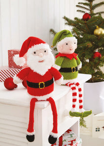 Hand knitted Santa and elf shelf sitter sitting on a chest of drawers with a Christmas tree behind them. Shelf sitters are toys with extra long legs. Santa is dressed in red and white, the elf is green with red and white striped legs