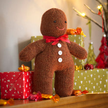 Load image into Gallery viewer, Image of a large knitted toy gingerbread man standing on a fireplace decorated with Christmas gifts and sweets. Knitted in a brown yarn with a red neck ribbon, 3 white buttons down the front, black eyes and an embroidered pink mouth