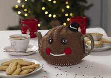 Load image into Gallery viewer, Gingerbread man tea cosy shown on a teapot on a festive table with a plate of gingerbread men biscuits. knitted in brown yarn, topped with a mini top hat. The facial features are black button eyes, a white embroidered mouth and rosy red cheeks