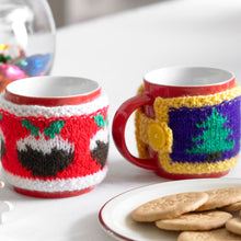 Load image into Gallery viewer, Two festive mug cosies shown on mugs next to a plate of biscuits. The first cosy is red with a white band top and bottom. Around the middle are Christmas puddings. The other has a purple band edged in yellow with Christmas trees around the centre.