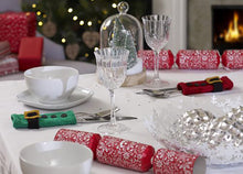 Load image into Gallery viewer, A festive table set for Christmas dinner and showing 2 hand knitted cutlery holders. A rectangle in stocking stitch with garter stitch top and bottom, a black belt, gold buckle and 3 black buttons. One is green for an elf and one is red for Santa