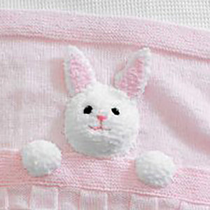 Cropped image of fake fur knitted rabbit head with black embroidered eyes and bright pink nose and mouth. Its ears have pink inner sections. You can also see its two front paws. It is sewn to the blanket to look like a rabbit in bed under a blanket