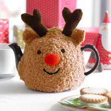 Load image into Gallery viewer, Rudolph the red nosed reindeer tea cosy displayed on a festive table with mince pies. Knitted in a light brown fur effect yarn with dark brown antlers in DK yarn. It is finished with a red pom pom nose and black embroidered eyes and mouth