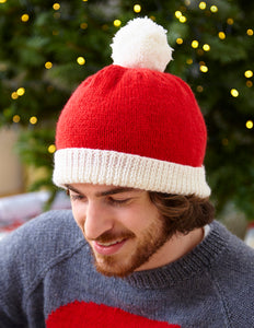 Man wearing a red and white beanie hat. The main section is knitted in stocking stitch with bright red DK yarn. The band is a white turn back knitted in rib. The hat is topped with a white pompom 