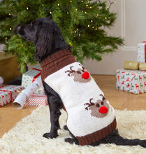 Load image into Gallery viewer, Black spaniel sitting wearing a festive coat hand knitted in cream DK yarn with brown collar and bans. On the back of the coat are two reindeer motifs knitted in light brown yarn with dark brown eyes and antlers. They are finished with a red pom pom nose
