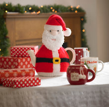 Load image into Gallery viewer, Image of a festive tea cosy displayed on a festive table with gift boxes. This Father Christmas tea cosy has a furry beard and traditional red and white hat and coat. He is finished off with a black belt and gold buckle