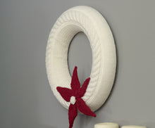 Load image into Gallery viewer, A simple white knitted wreath with a cable twist running around the front. A five pointed poinsettia is knitted in red and attached to the bottom front with a white bobble at the centre