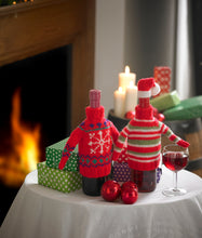 Load image into Gallery viewer, 2 wine bottle jumpers sitting on a festive decorated table. One is red with a Fair Isle design - white snowflake and green and blue details. The other is red, white and green stripes with red bands and neck, topped with a white brimmed, red Santa hat