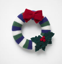 Load image into Gallery viewer, Christmas wreath hand knitted in royal blue, dark green and white stripes in garter stitch. Hand knitted holly leaves with red berries are attached to the bottom right and, at the top, is a rich red bow knitted in garter stitch