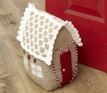 Load image into Gallery viewer, A knitted gingerbread doorstop. Knitted in light brown yarn. On the side are 2 windows each made of 4 white panes of 'glass'. The red door is outlined with a red and white twisted yarn in the shape of a heart. The white latticed roof has a picot edge