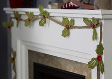 Load image into Gallery viewer, A holly garland pictured on a mantlepiece. A brown knitted ribbon forms the 'string' and placed at intervals are 3 green holly leaves with a red button sewn at their centre to attach the to the ribbon