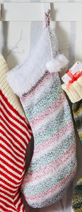 Image of smaller size Christmas stocking hand knitted in self striping super chunky yarn. The stripes are pink, green, grey and white and the top is knitted with white tinsel for a sparkly finish
