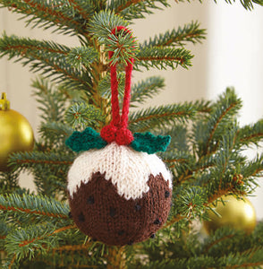 A Christmas pudding bauble hanging on a Christmas tree. Knitted in dark brown yarn with black 'dots'. The top is white yarn knitted to look like the icing or cream is running down. Topped with green holly leaves, red berries and a red hanging loop