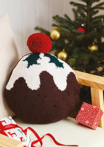 Fun Christmas pudding cushion shown on a grey armchair. The main section is circular and knitted in dark brown DK yarn. The white top is knitted to look like the topping is running down the pudding. Finished with holly leaves and a big red pom pom
