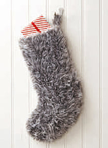 Image of a large Christmas stocking hand knitted in grey or silver coloured faux fur yarn. Pictured hanging on a red door with a wrapped present sticking out the top