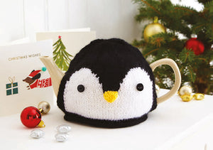 Penguin tea cosy sitting on a festive table. Hand knitted in black yarn with a white front. Finished with black eyes and a yellow beak
