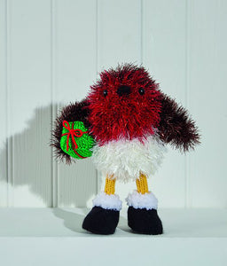 Robin toy knitted in tinsel yarn. The wings are brown. The main front section is dark red and the bottom of the body is white. He has yellow stick legs and black boots in DK yarn. The boots have white fur tops. Holds a green gift with red bow