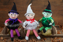 Load image into Gallery viewer, 3 hand-knitted witches sitting on wooden pumpkins. They all have a hat, neck tie, tinsel body and striped stockings and are holding a broomstick. One is black with pink arms, one is white and silver with red and white stockings and the other is green
