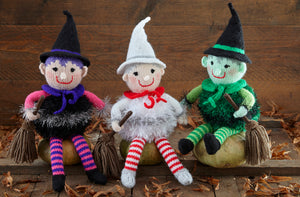 3 hand-knitted witches sitting on wooden pumpkins. They all have a hat, neck tie, tinsel body and striped stockings and are holding a broomstick. One is black with pink arms, one is white and silver with red and white stockings and the other is green