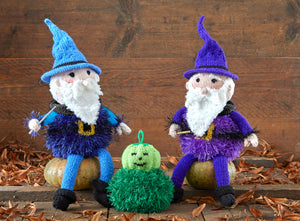 Image of 2 hand knitted wizards sitting on wooden pumpkins with a green mini knitted pumpkin on the ground between them. 1 wizard has a blue hat, arms and legs and a dark blue tinsel body, the other is knitted in shades of purple