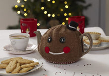 Load image into Gallery viewer, Gingerbread man tea cosy shown on a teapot on a festive table with a plate of gingerbread men biscuits. knitted in brown yarn, topped with a mini top hat. The facial features are black button eyes, a white embroidered mouth and rosy red cheeks

