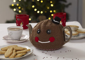 Gingerbread man tea cosy shown on a teapot on a festive table with a plate of gingerbread men biscuits. knitted in brown yarn, topped with a mini top hat. The facial features are black button eyes, a white embroidered mouth and rosy red cheeks