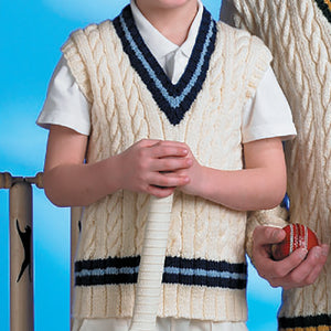 Cropped image of young boy holding a cricket bat and wearing a traditional cable hand knit cricket slipover or vest. The vest has a v neck and is knitted in cream yarn with black and blue stripe round the bottom and for the V neck