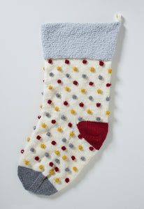 Image of a large festive holiday stocking with a light silver fur texture band and a hanging loop. Knitted in cream DK yarn with a dark red heel and dark grey toe piece. The bobbles are all over the cream section and are knitted grey, red and mustard