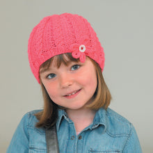 Load image into Gallery viewer, Image of young girl wearing a hand knitted Aran beanie in pink yarn. The hat has a short rib band and the main section is knitted with alternating reverse rib and cable twists. A pink flower to attached to the front with a white button
