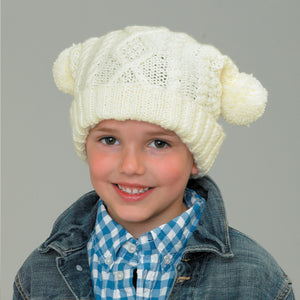 Image of young boy wearing a hand knitted Aran tea bag hat in cream or off-white yarn. The hat has a cable diamond panel at the front and stripes of cable twists. It is finished with two pom poms which fold over at the corners and a turn back in rib