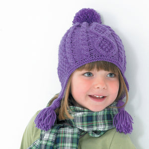Image of young girl wearing a hand knitted Aran hat in purple yarn. The hat is a pom pom or bobble helmet style hat with ear flaps and tassels. The main section is knitted in moss or seed stitch with cable twist and a diamond cable panel at the front