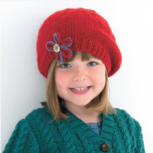 Load image into Gallery viewer, Image of young girl wearing a hand knitted Aran beret in red yarn. A super cute beret design with rib band and a blue and red flower added to the front with a button in the centre 

