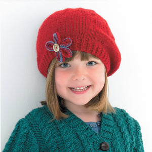 Image of young girl wearing a hand knitted Aran beret in red yarn. A super cute beret design with rib band and a blue and red flower added to the front with a button in the centre 