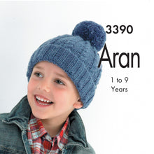 Load image into Gallery viewer, Image of young boy smiling and wearing a hand knitted Aran hat in denim blue yarn. The hat is a pom pom or bobble hat with rib turn back and cable twists on the main section
