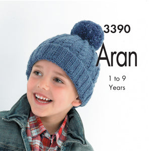 Image of young boy smiling and wearing a hand knitted Aran hat in denim blue yarn. The hat is a pom pom or bobble hat with rib turn back and cable twists on the main section