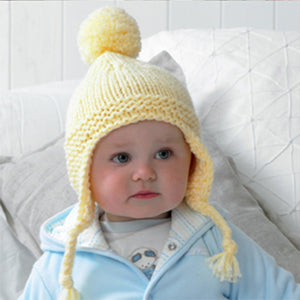 The baby in this image is wearing a hand knitted chunky baby hat with ear flaps and a pom pom. This bobble hat is knitted with yellow yarn and has a garter stitch edge and ear flaps. The flaps are finished with braids that are knotted at the end