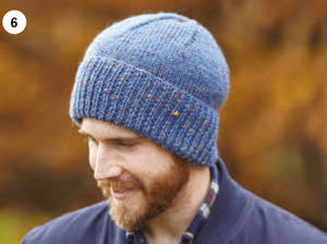 Image of a man wearing a snug fitting beanie hat. The hat is hand knitted in a blue Aran yarn with light colourful flecks. The hat features a deep rib turn back and the main section is knitted in plain stocking stitch