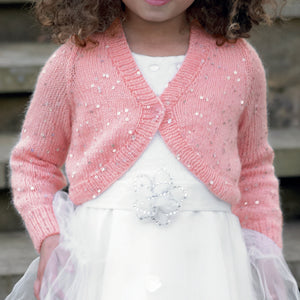 Close up image of young girl wearing a white dress and pink v neck bolero cardigan or shrug. The bolero has long sleeves and the yarn is interwoven with silver sequins to make it sparkle