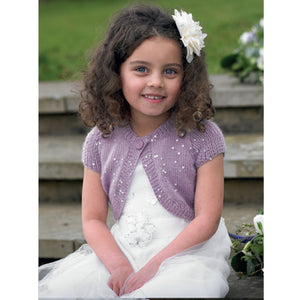 Image of young girl wearing a white dress with a white flower in her hair. She is also wearing a dusky purple round neck bolero cardigan or shrug. The bolero has capped sleeves and the yarn is interwoven with silver sequins to make it sparkle