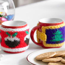 Load image into Gallery viewer, Two festive mug cosies shown on mugs next to a plate of biscuits. The first cosy is red with a white band top and bottom. Around the middle are Christmas puddings. The other has a purple band edged in yellow with Christmas trees around the centre.
