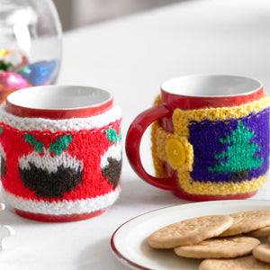 Two festive mug cosies shown on mugs next to a plate of biscuits. The first cosy is red with a white band top and bottom. Around the middle are Christmas puddings. The other has a purple band edged in yellow with Christmas trees around the centre.