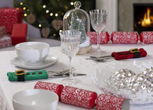 Load image into Gallery viewer, A festive table set for Christmas dinner and showing 2 hand knitted cutlery holders. A rectangle in stocking stitch with garter stitch top and bottom, a black belt, gold buckle and 3 black buttons. One is green for an elf and one is red for Santa
