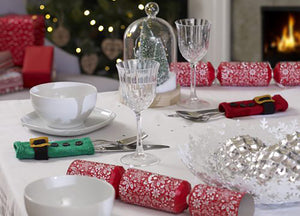 A festive table set for Christmas dinner and showing 2 hand knitted cutlery holders. A rectangle in stocking stitch with garter stitch top and bottom, a black belt, gold buckle and 3 black buttons. One is green for an elf and one is red for Santa