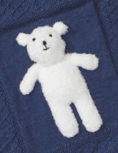 Close up of a white teddy bear hand knitted in fake fur yarn. It has black embroidered face features and is attached to a navy baby blanket