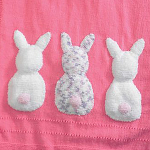 Crop of the 3 rabbit backs sewn in a row at the bottom of the plain blanket. The left and right rabbits are knitted in white faux fur with a pale pink pompom tail. The middle rabbit is knitted in white with purple and pink flecks and a pale pink tail