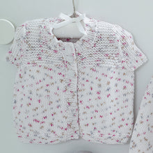 Load image into Gallery viewer, Image of a short sleeve cardigan hanging on a hanger. The tops of the fronts are knitted in garter stitch and the rest is knitted in stocking stitch. The yarn is a white cotton DK yarn with white and silver flecks

