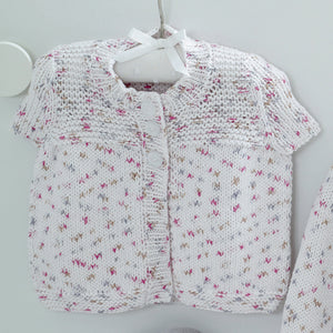 Image of a short sleeve cardigan hanging on a hanger. The tops of the fronts are knitted in garter stitch and the rest is knitted in stocking stitch. The yarn is a white cotton DK yarn with white and silver flecks